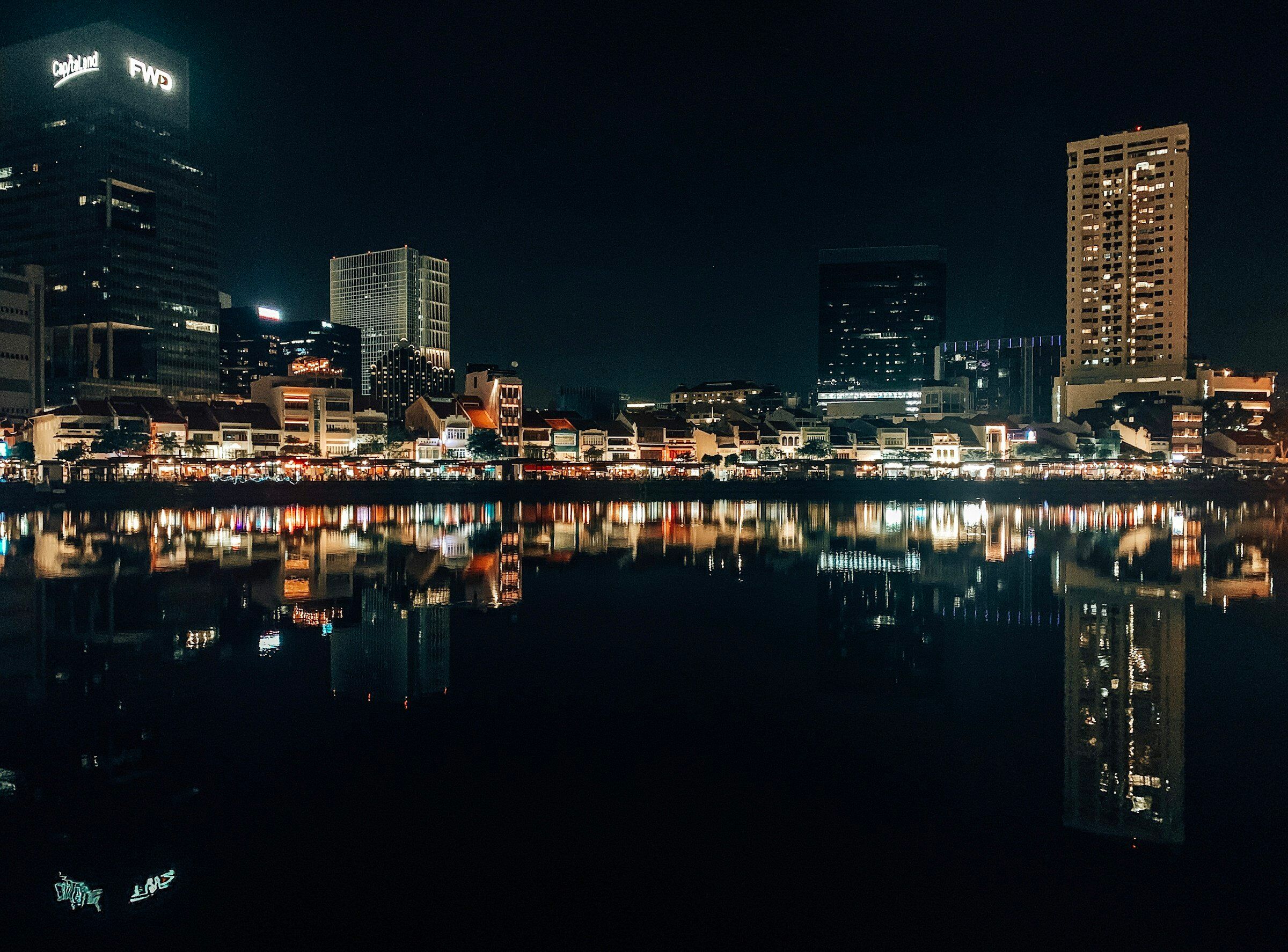 Boat Quay's shophouse row lit at night across the Singapore River, the lamps reflected in the water